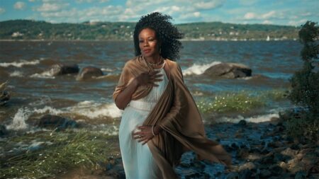 Artist/activist Dragonfly (Robin LaVerne Wilson) standing gracefully on the sandy banks of the shore, as a Black woman in a Grecian white robe who embodies the mythic Aphrodite, posing as portrayed in Botticelli’s “Birth of Venus”