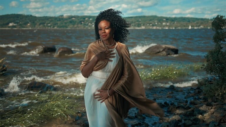 Artist/activist Dragonfly (Robin LaVerne Wilson) standing gracefully on the sandy banks of the shore, as a Black woman in a Grecian white robe who embodies the mythic Aphrodite, posing as portrayed in Botticelli’s “Birth of Venus”